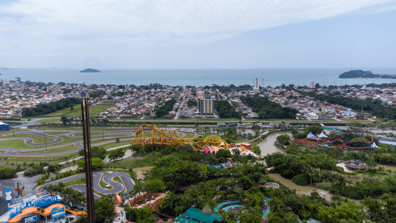 Vista aérea de um parque temático com montanhas-russas amarelas, árvores exuberantes e uma cidade costeira com praia ao fundo.