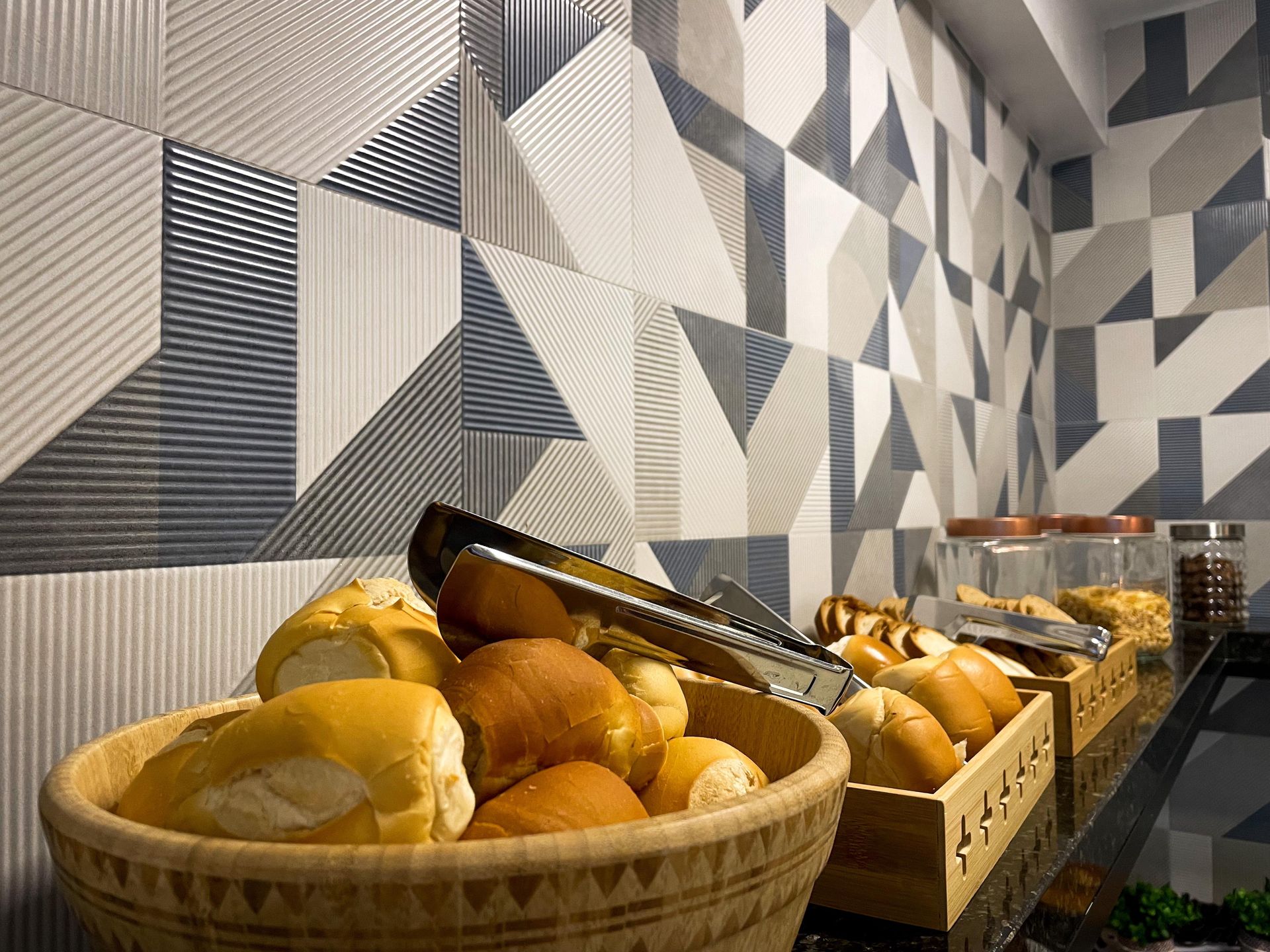 Duas cestas de madeira cheias de pãezinhos recém-assados ​​estão sobre uma superfície encostada em uma parede de azulejos com padrões geométricos.