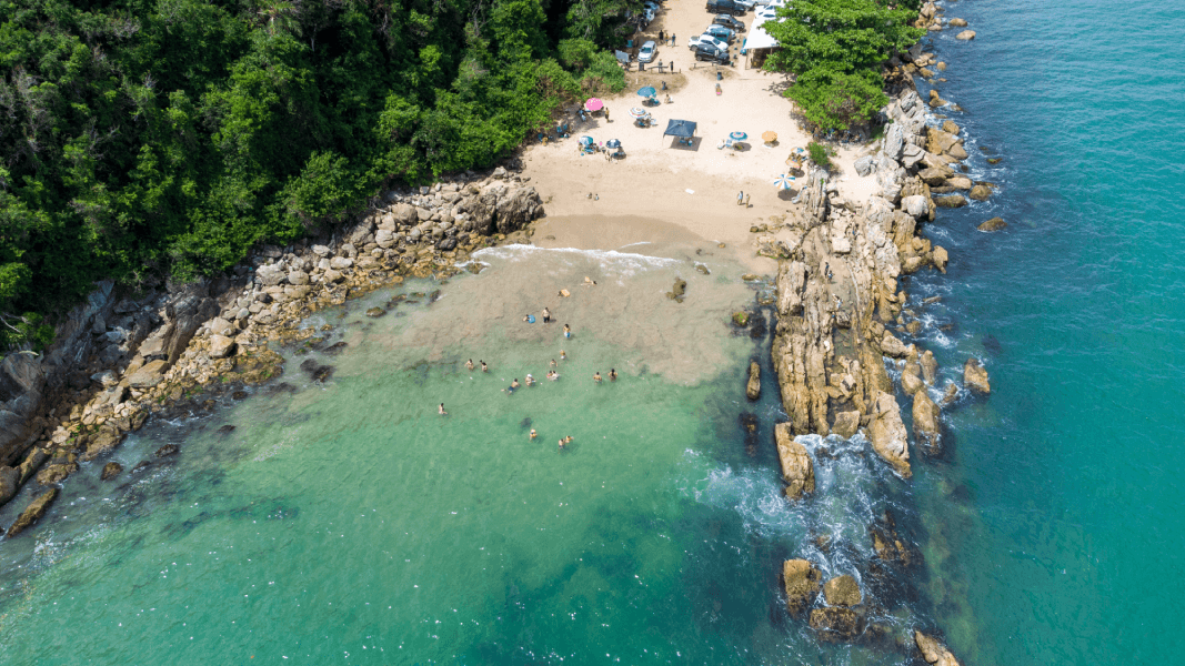 Vista aérea de uma pequena enseada de areia com água turquesa, margens rochosas e densa floresta margeando a praia.