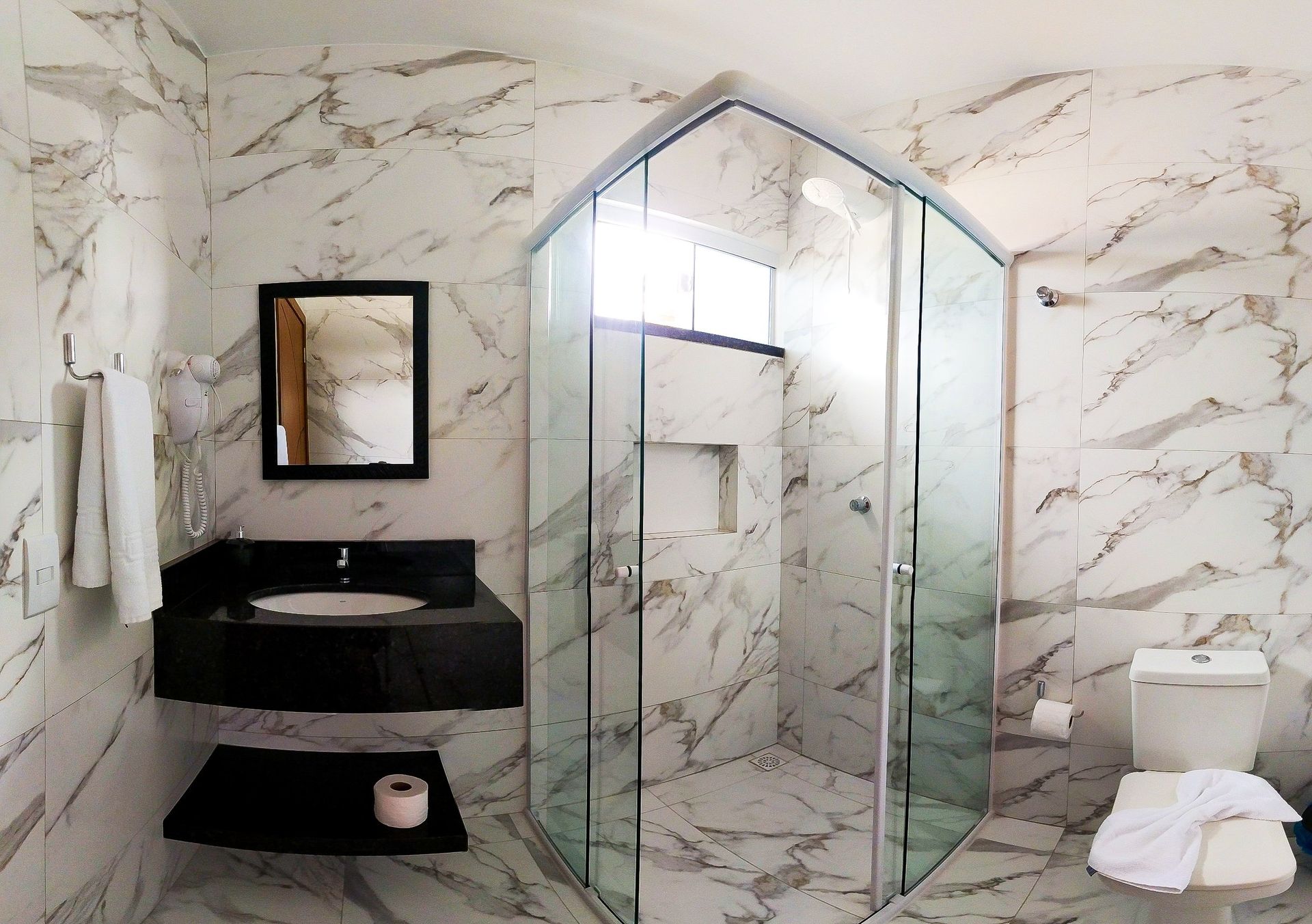 Um banheiro com azulejos de mármore, box de vidro para o chuveiro, pia preta com armário e vaso sanitário branco.