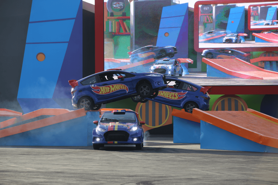 Carrinhos de acrobacias Hot Wheels azuis realizam um salto sincronizado no ar em uma pista de obstáculos colorida com tema de pista de corrida.