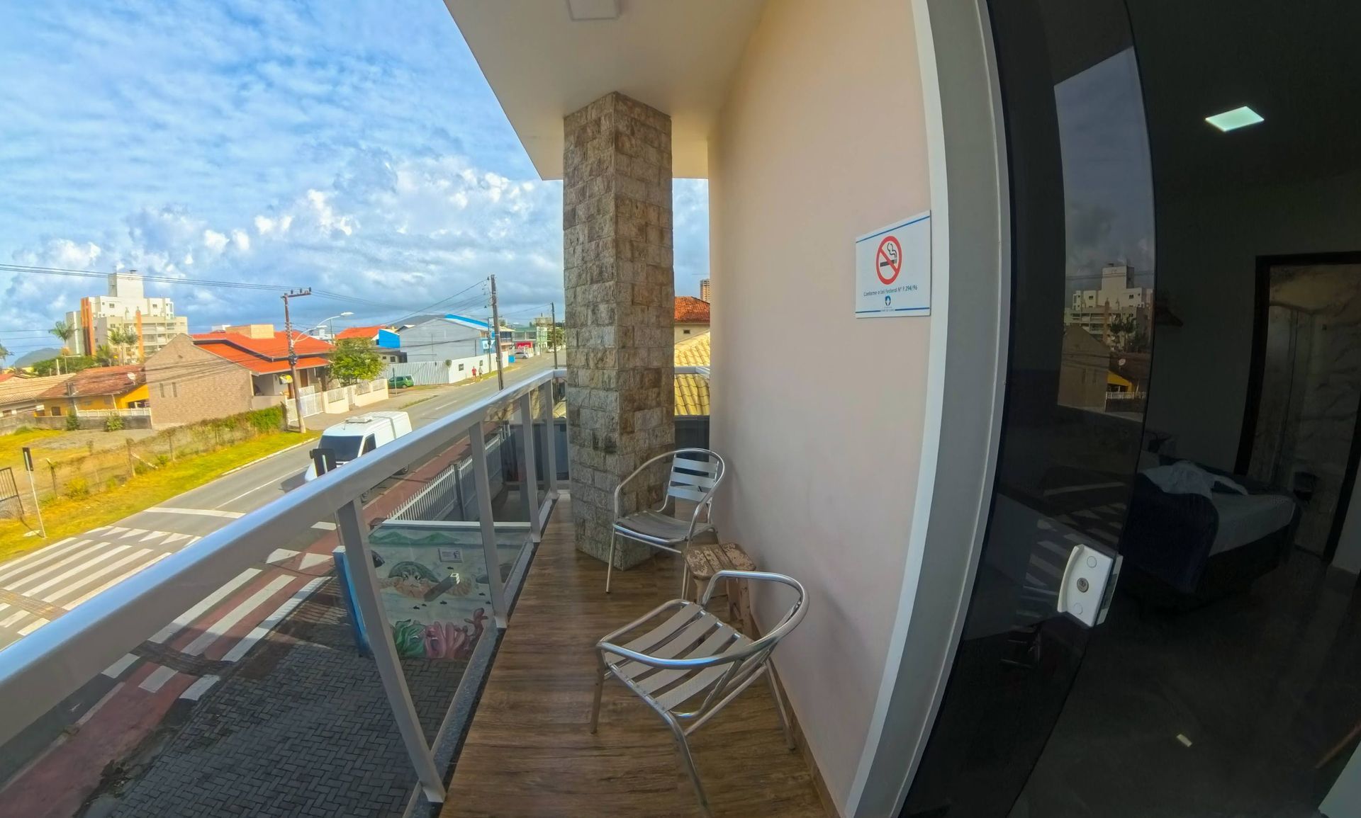 Uma varanda de hotel com duas cadeiras de metal, um pilar de pedra e vista para uma rua e prédios sob um céu nublado.