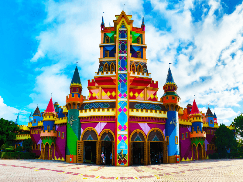 Um castelo colorido e extravagante, com torres altas e uma fachada vibrante com diversos padrões, contrastando com um céu azul e nublado.