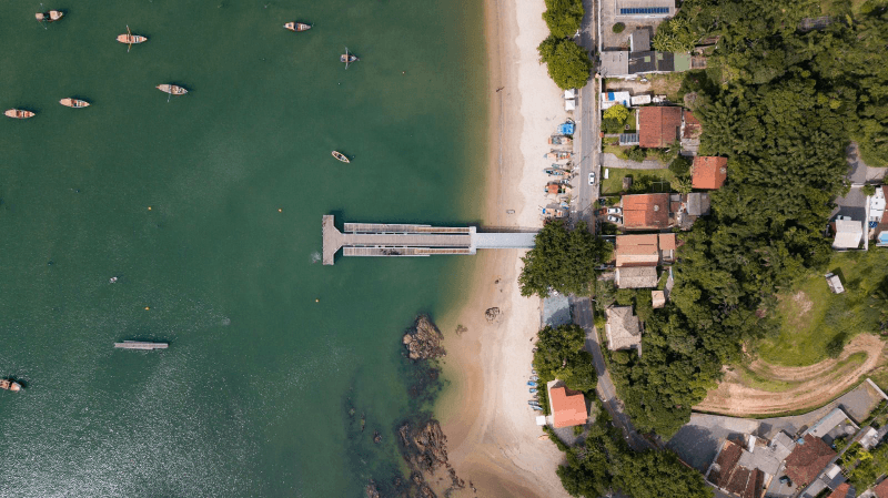 Vista aérea de um cais que se estende de uma praia de areia até um mar verde com pequenos barcos, ao lado de casas e árvores.