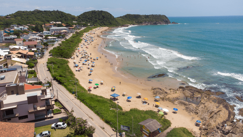 Vista aérea de uma praia de areia com guarda-sóis coloridos, ondas do oceano e casas costeiras aninhadas em uma encosta verdejante.