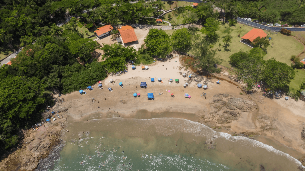 Vista aérea de uma pequena praia de areia situada numa enseada costeira exuberante e verdejante, com vários guarda-sóis coloridos.