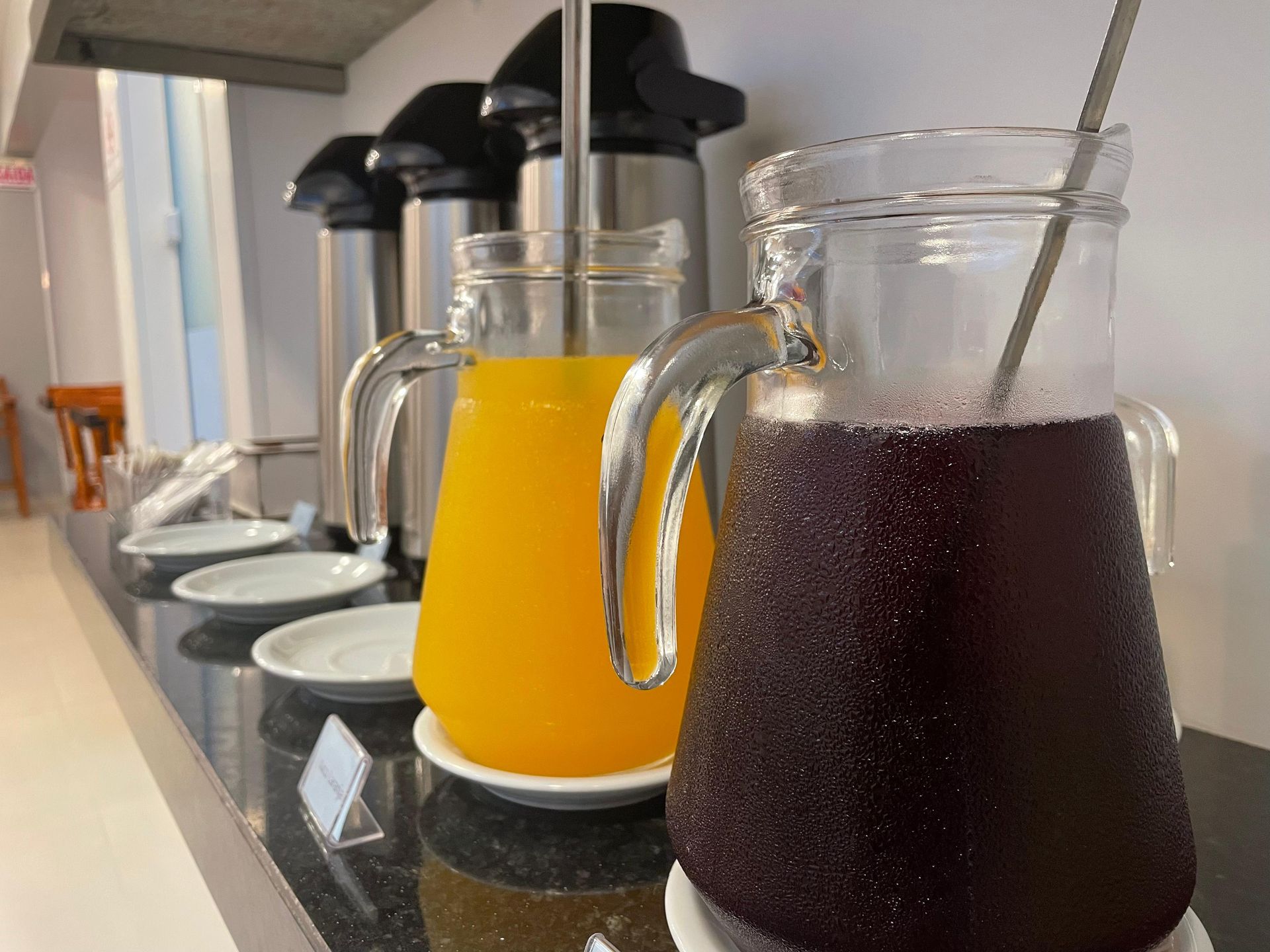 Duas jarras de vidro com suco, uma de laranja e outra roxa, estão sobre uma bancada ao lado de pratos empilhados e garrafas de café.
