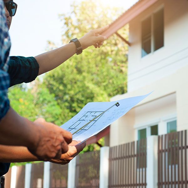 Person holding a blueprint while another points toward a sunlit house exterior