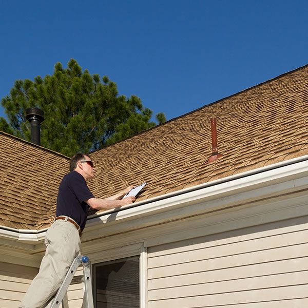 Man standing on a ladder inspecting a tan shingled roof with a clipboard under a clear blue sky