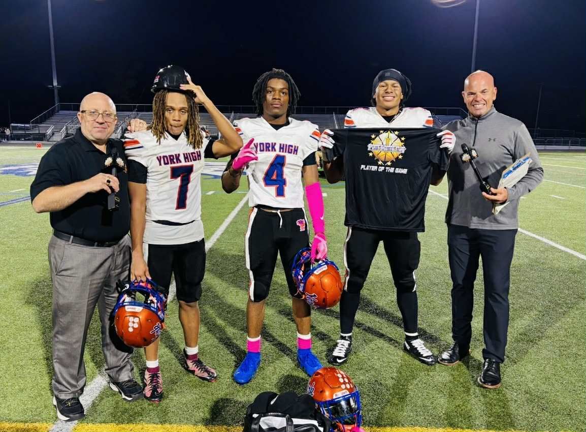 Five people stand on a football field at night holding jerseys and helmets, posing for a photo.