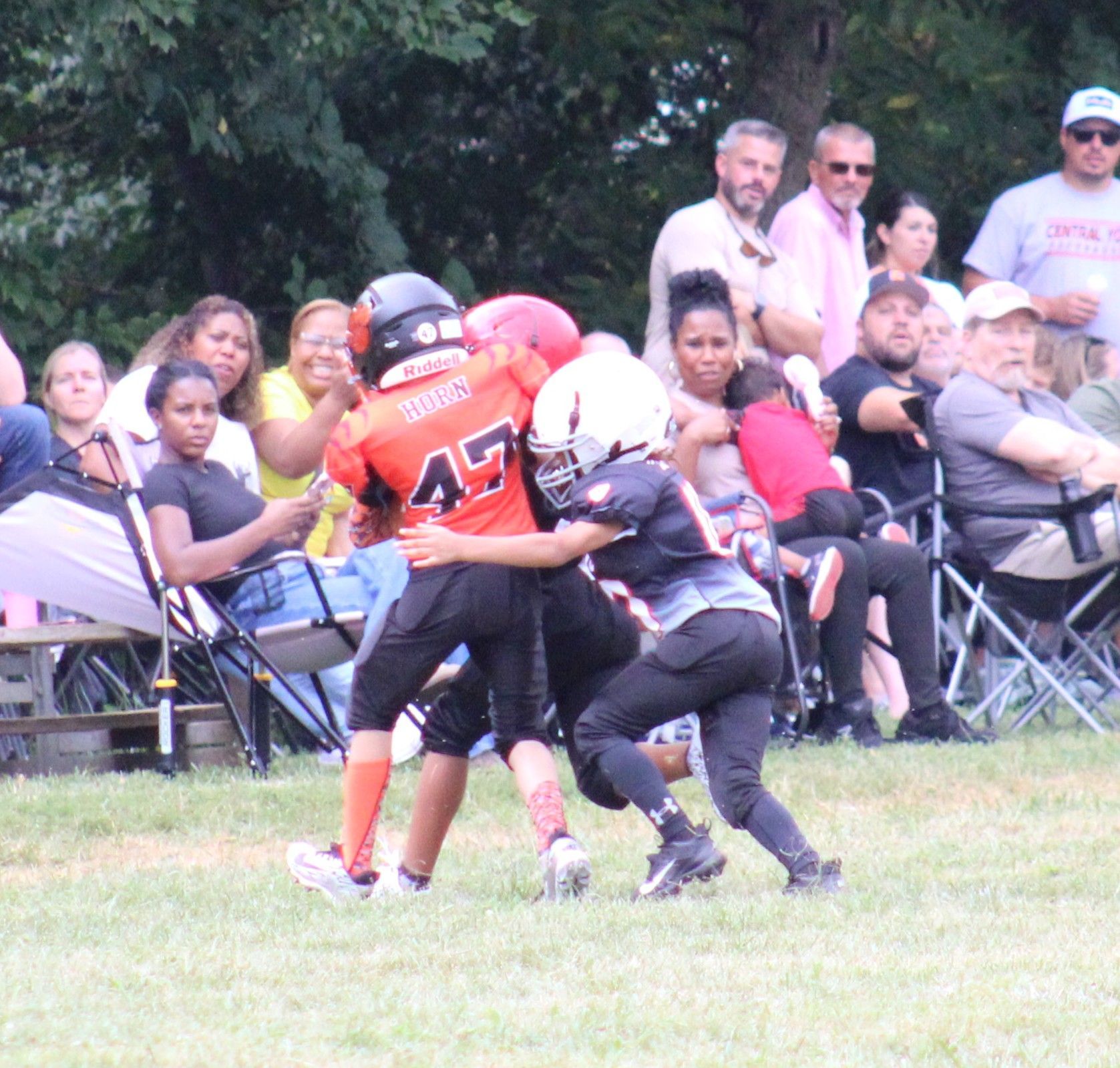 Two youth football players in red and black collide on a grassy field while spectators watch behind them