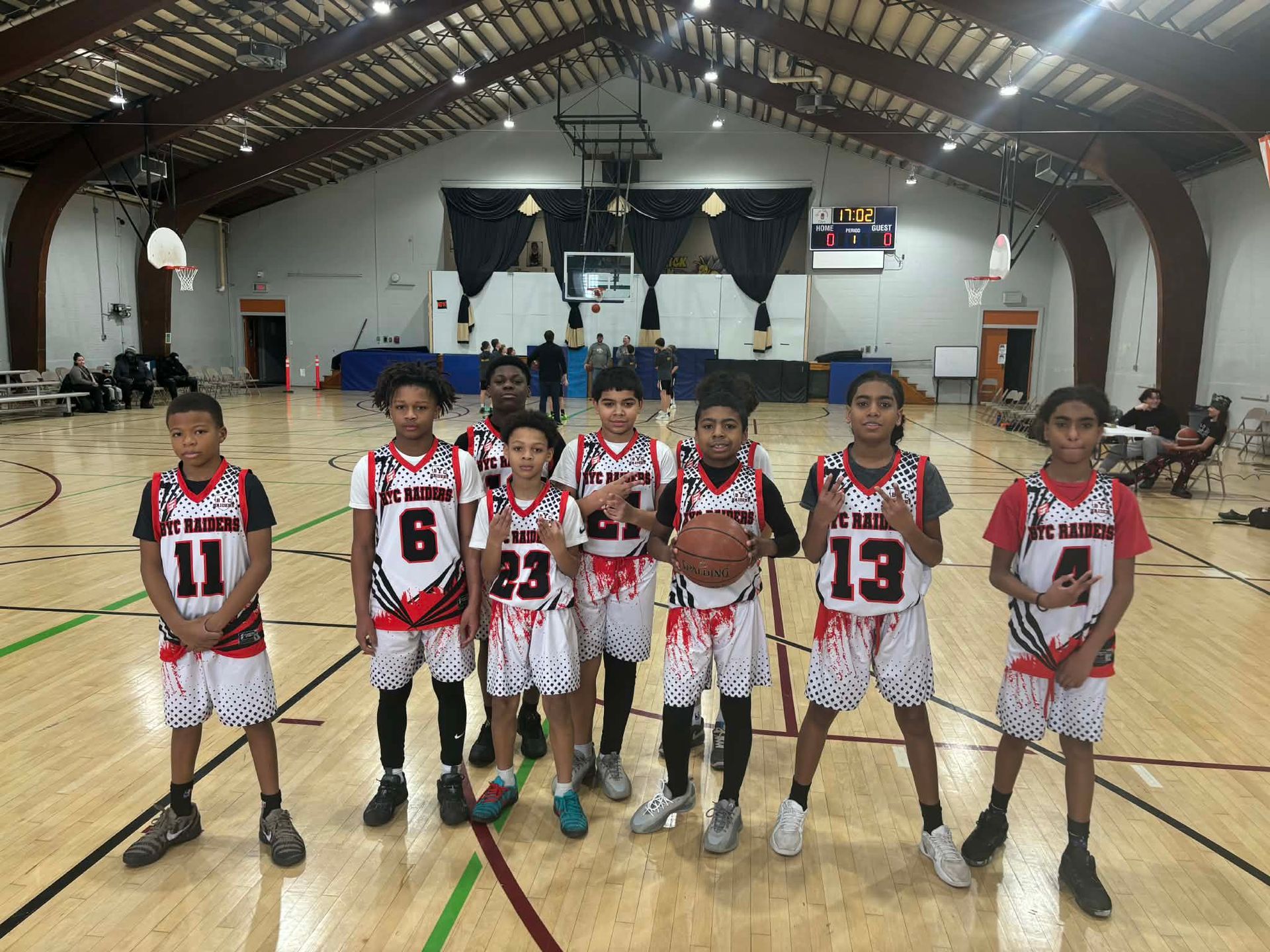 Youth basketball team posing on an indoor court, wearing red-and-white jerseys with numbers in front of the hoop.