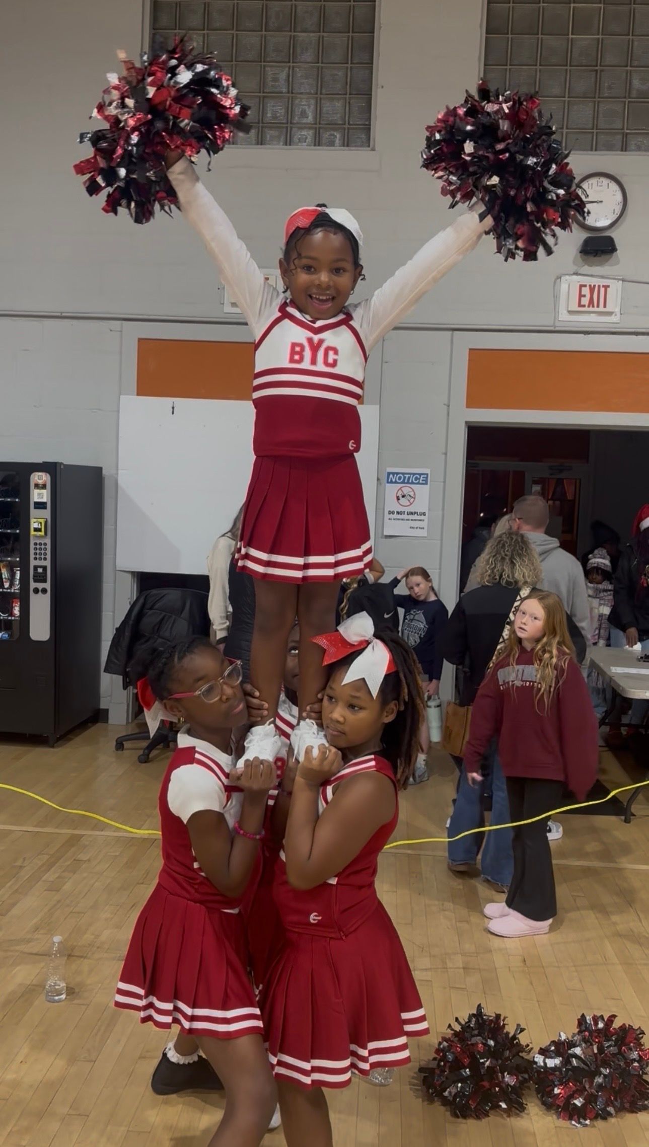 Cheerleaders in red-and-white uniforms pose in a gym, one lifted with pom-poms raised.