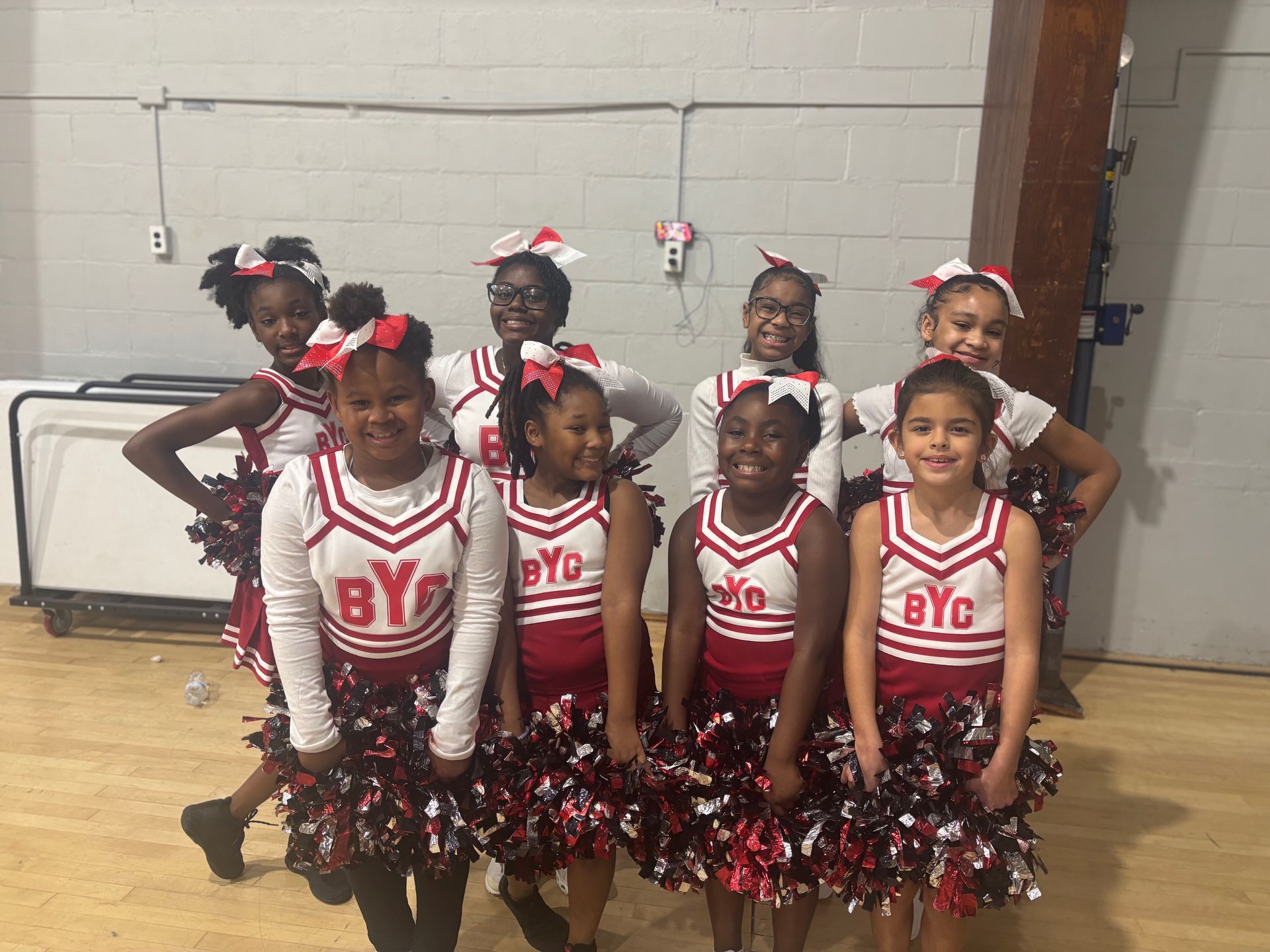 Cheer team in red-and-white uniforms posing indoors with pom-poms and hair bows