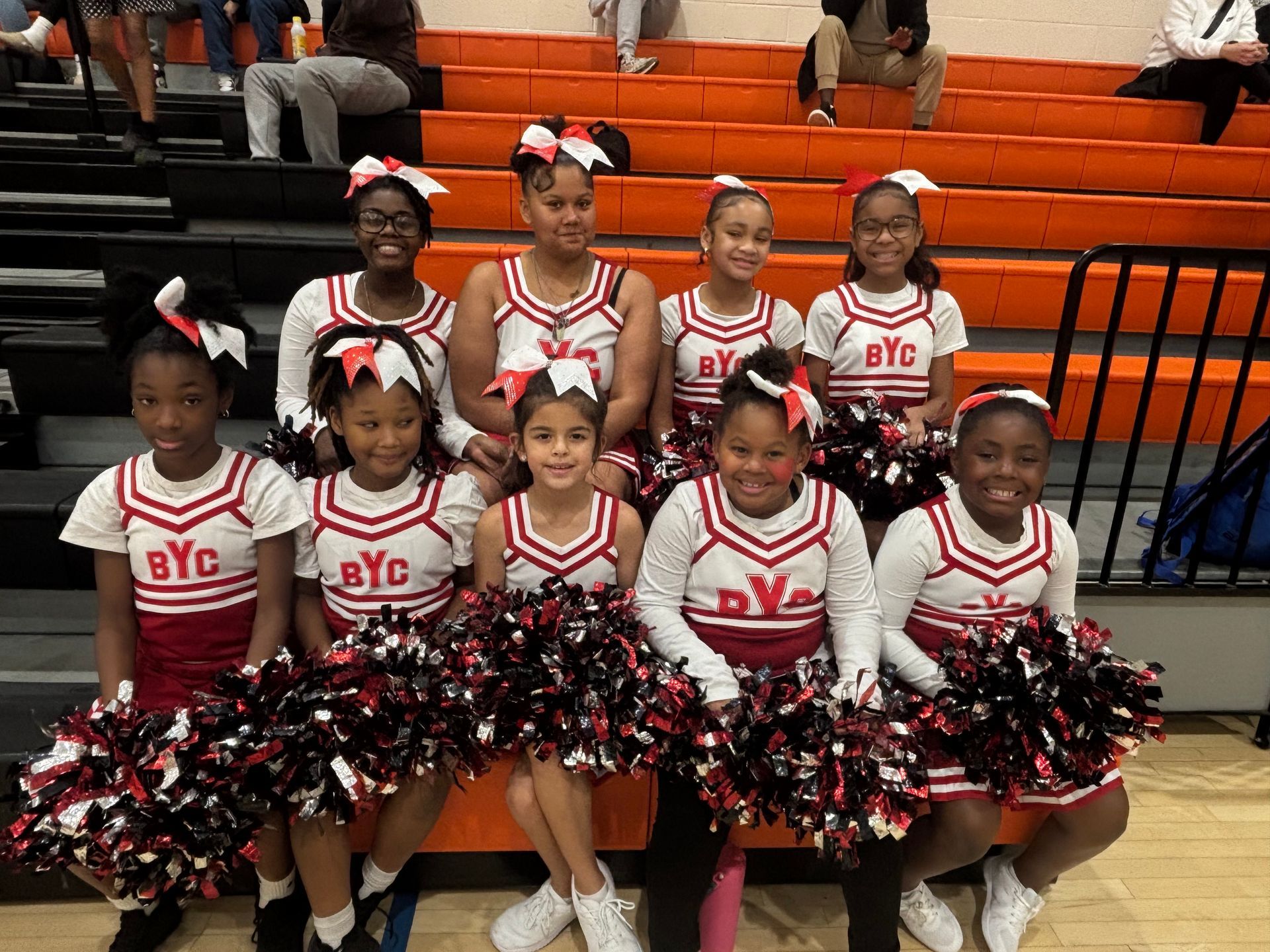 Cheer squad in white and red uniforms posing with black-and-red pom-poms on bleachers