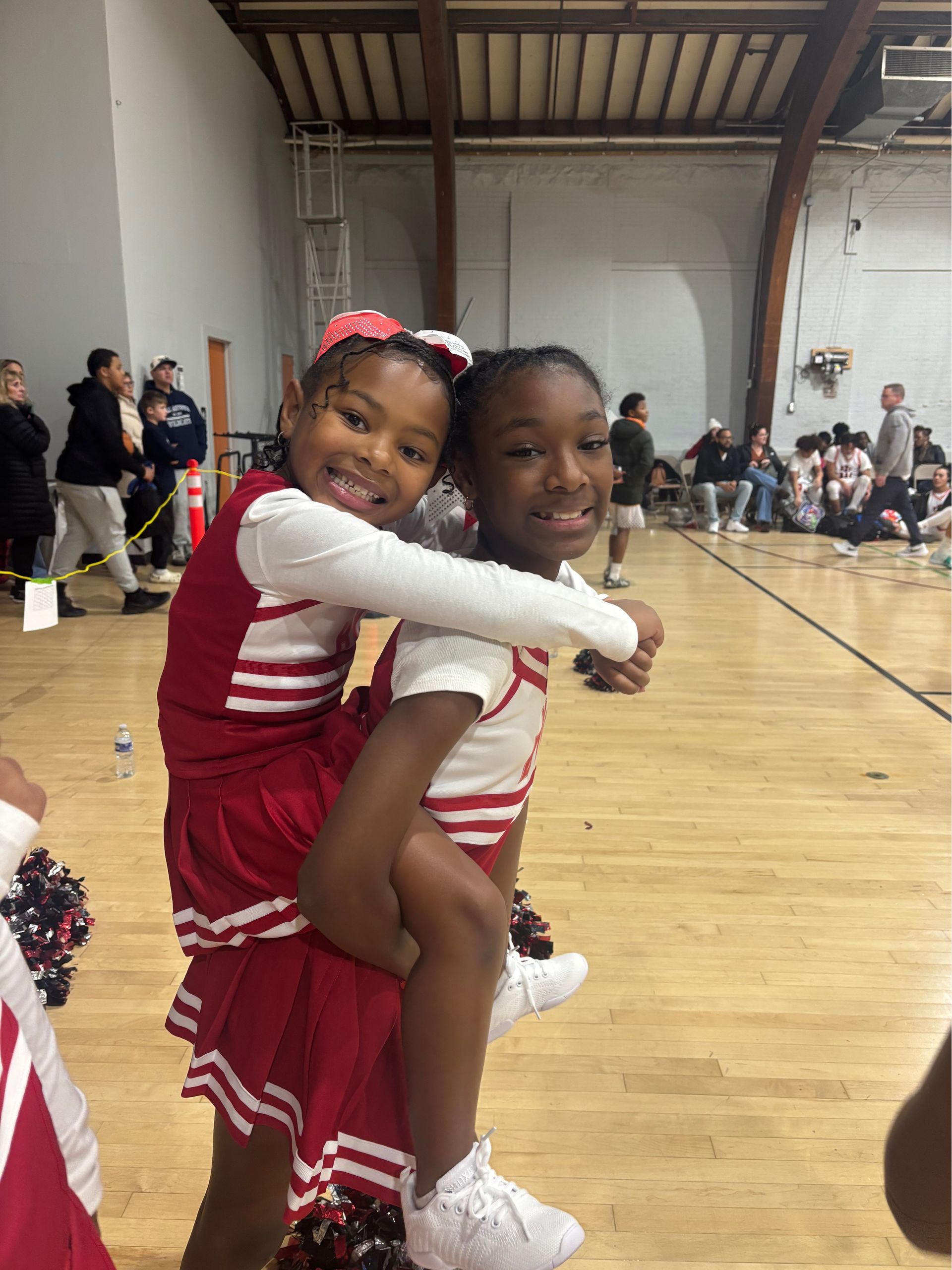 Two cheerleaders in red and white uniforms hugging in a gymnasium during a performance.