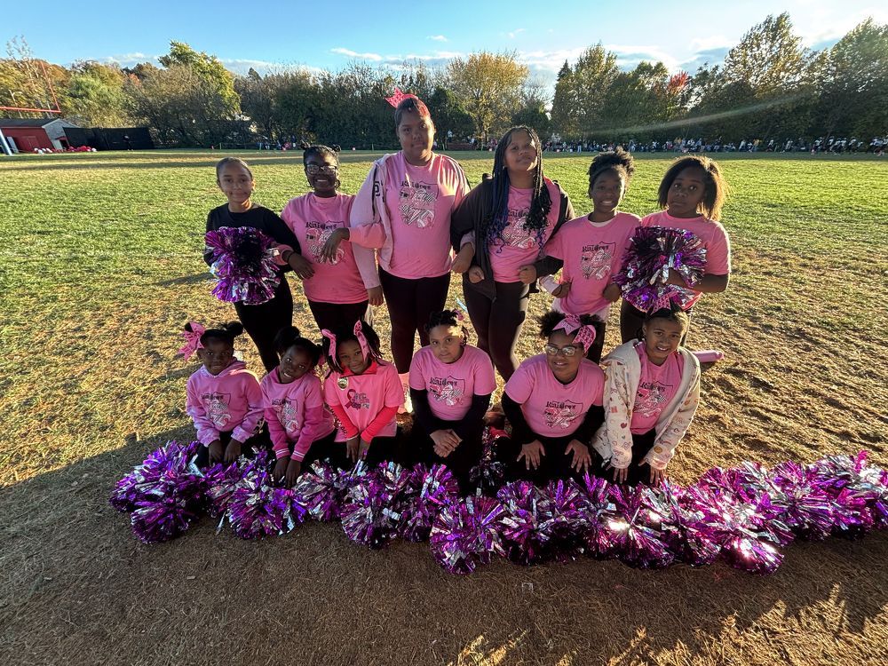 Youth sports team posing on a field with pink shirts and pom-poms, with trees in the background.