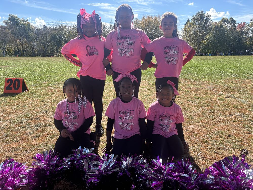 Six children in bright pink shirts pose on a grassy field with purple pom-poms in front.