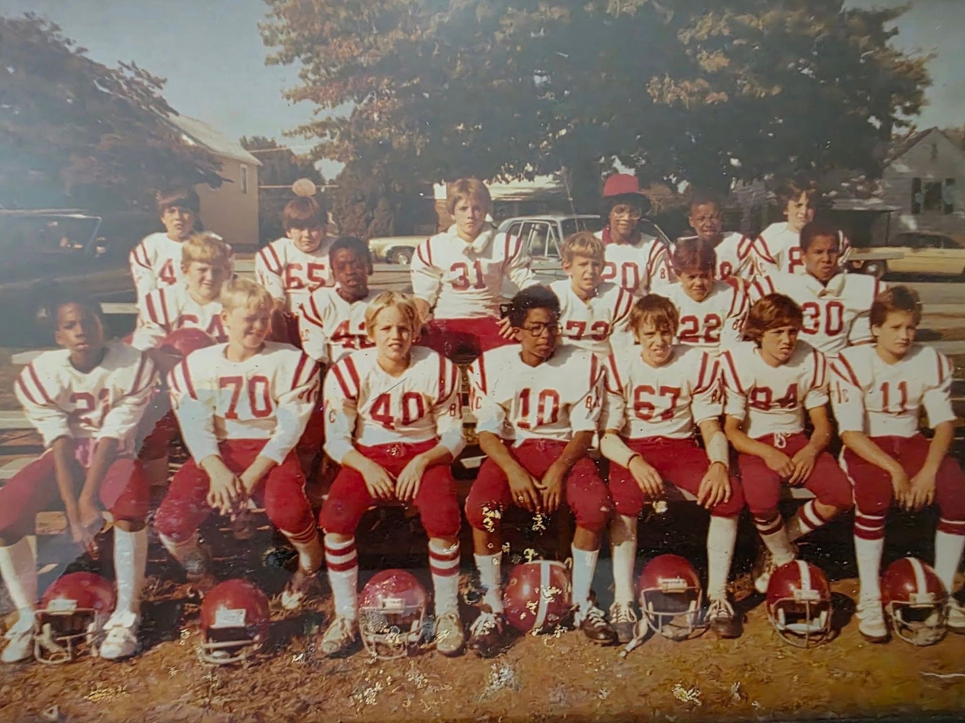 Youth football team posed in red-and-white uniforms, kneeling and standing outdoors with helmets in front.