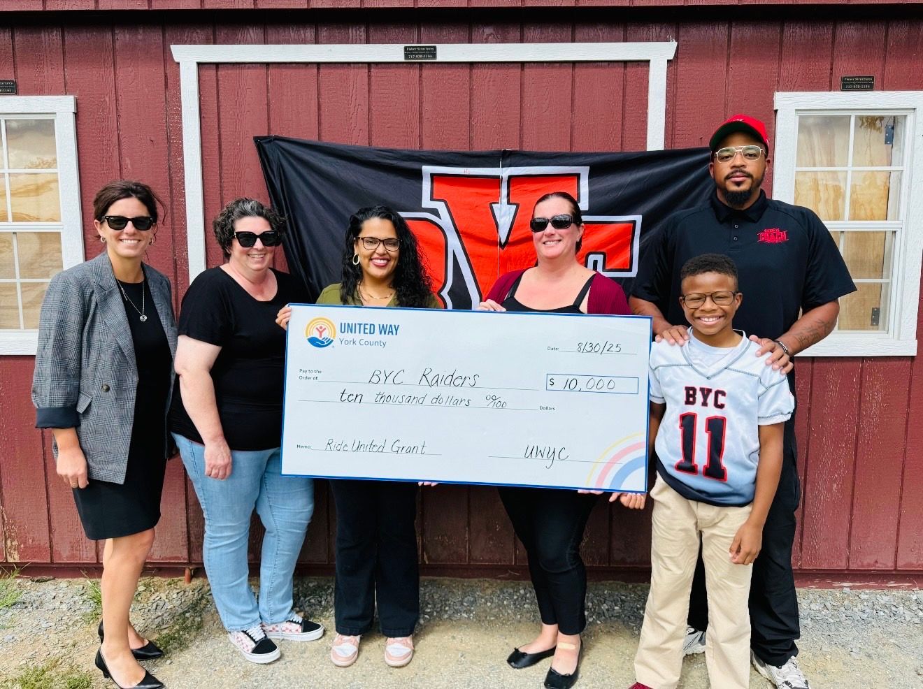 Group holding oversized check in front of red barn with black-and-red banner