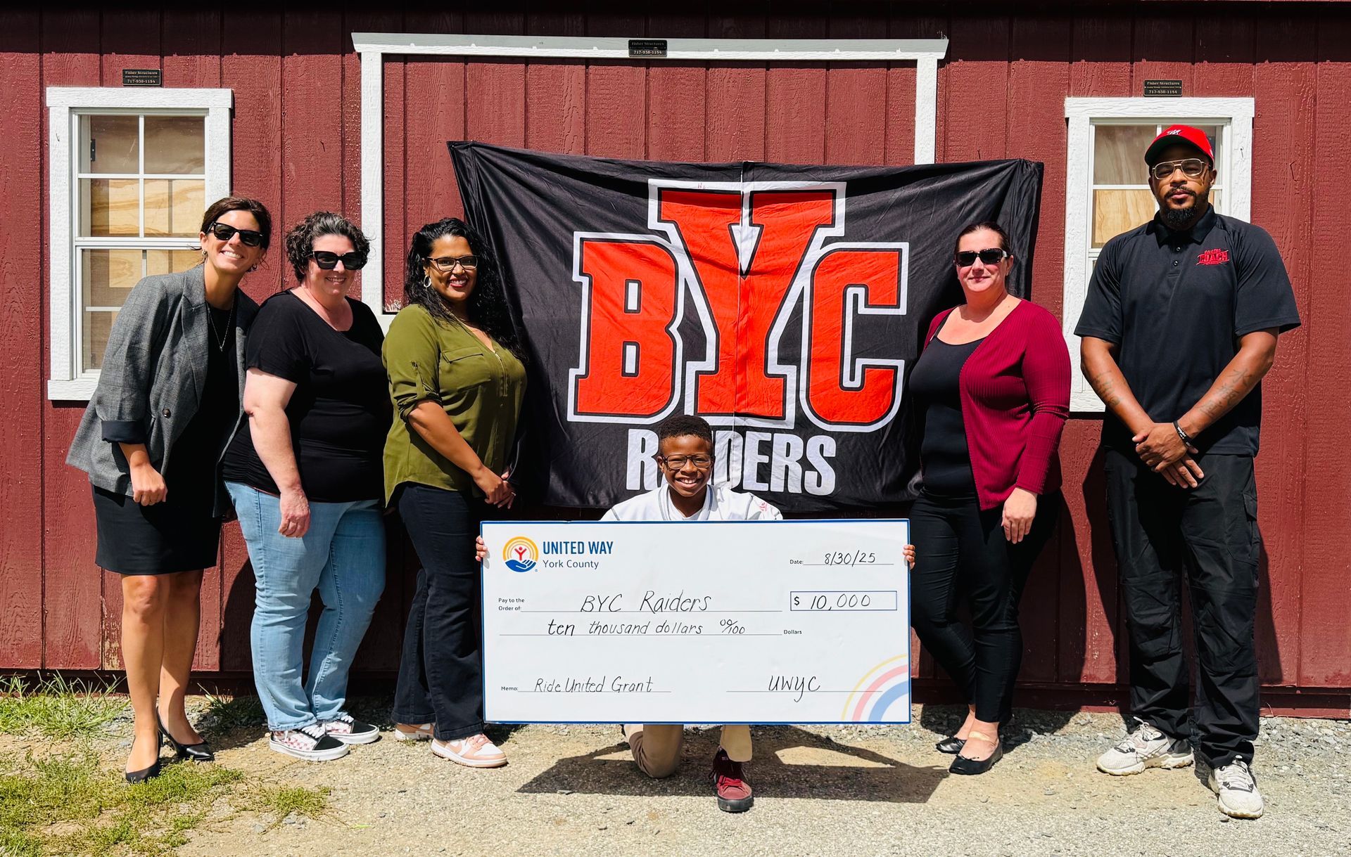 Group posing in front of a BVC Raiders banner, holding a large check outside a brick building