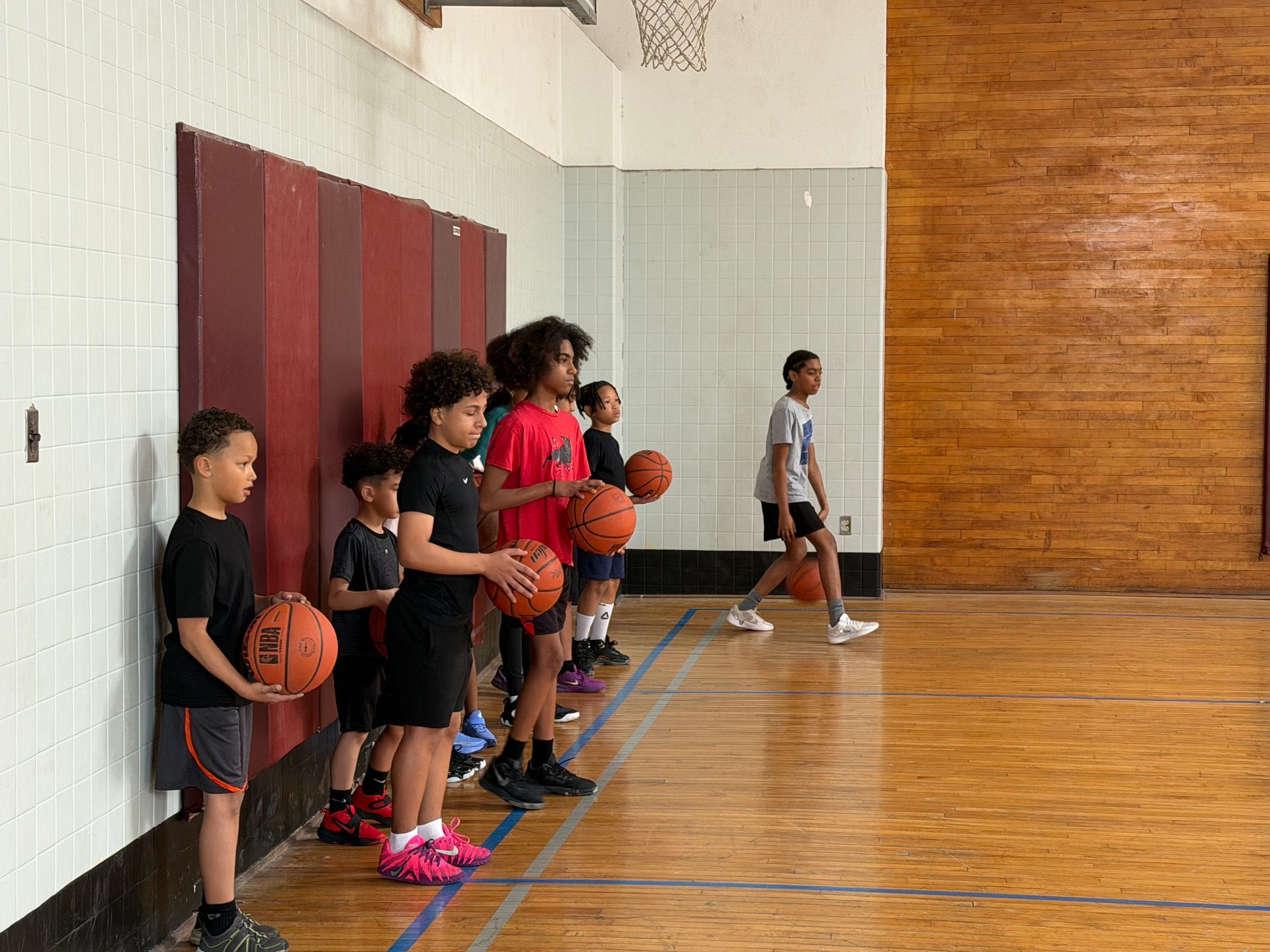 Children lined up in a gym holding basketballs near a court wall and hoop