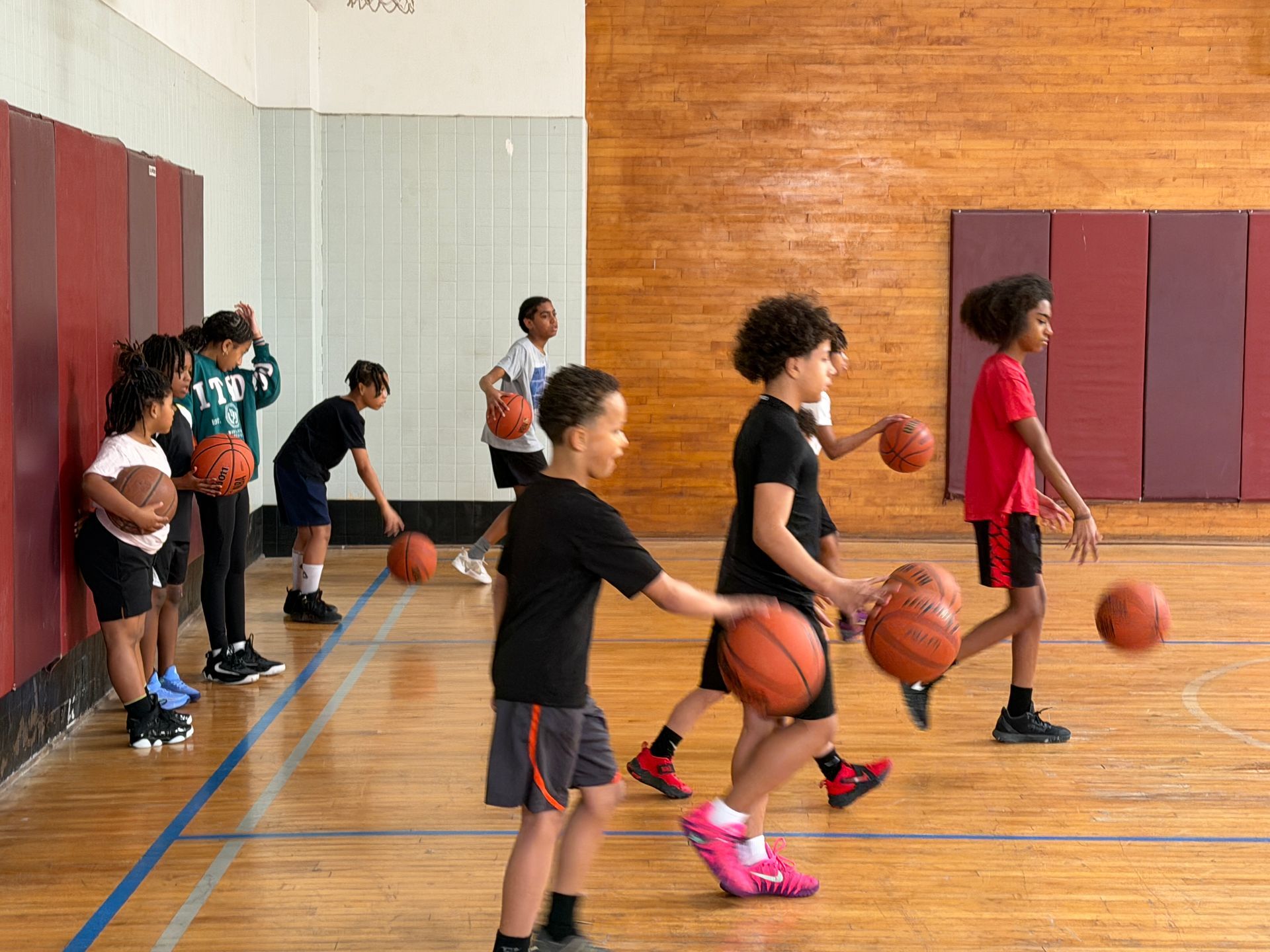 Children practicing basketball drills in a gym, lined up with balls near the wall.