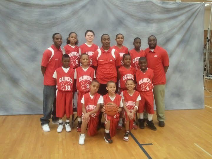 Youth basketball team posing in red uniforms in a gymnasium, with coaches standing behind them.