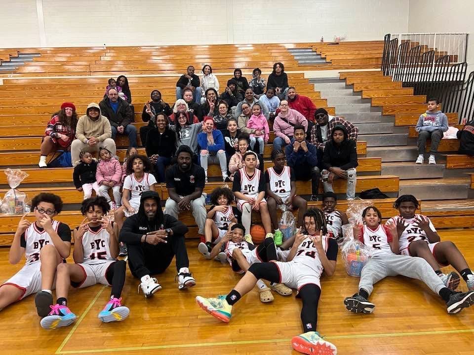 Basketball team and supporters posed on a gym bleacher after a game, with players seated in front.