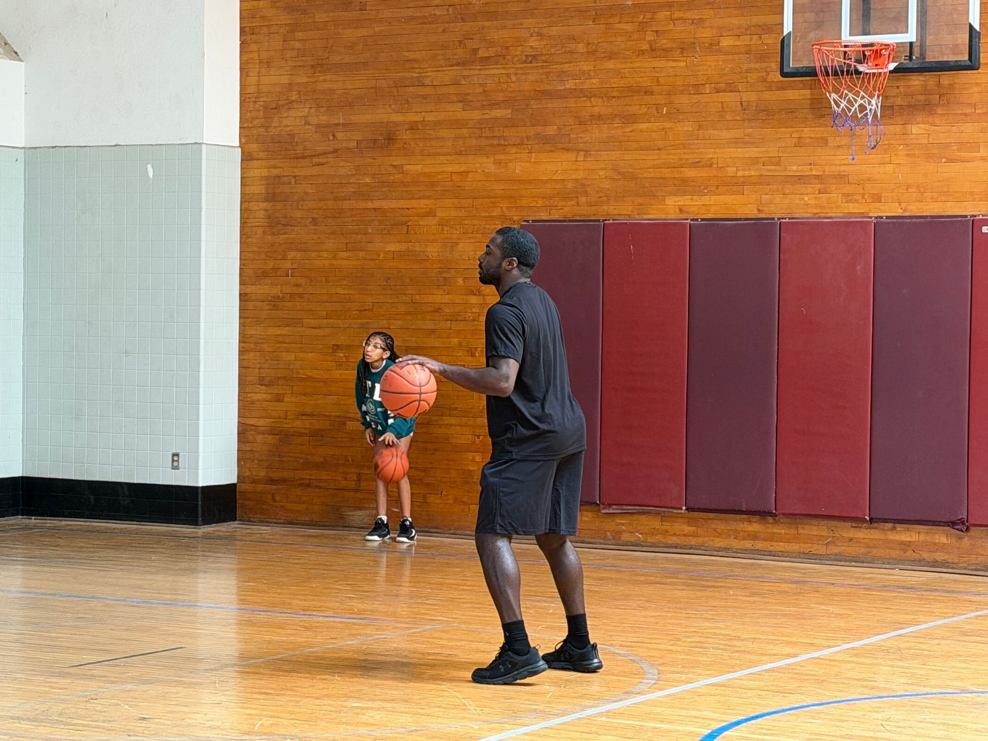 Man holding a basketball in a gym, facing a child near the wall and hoop.