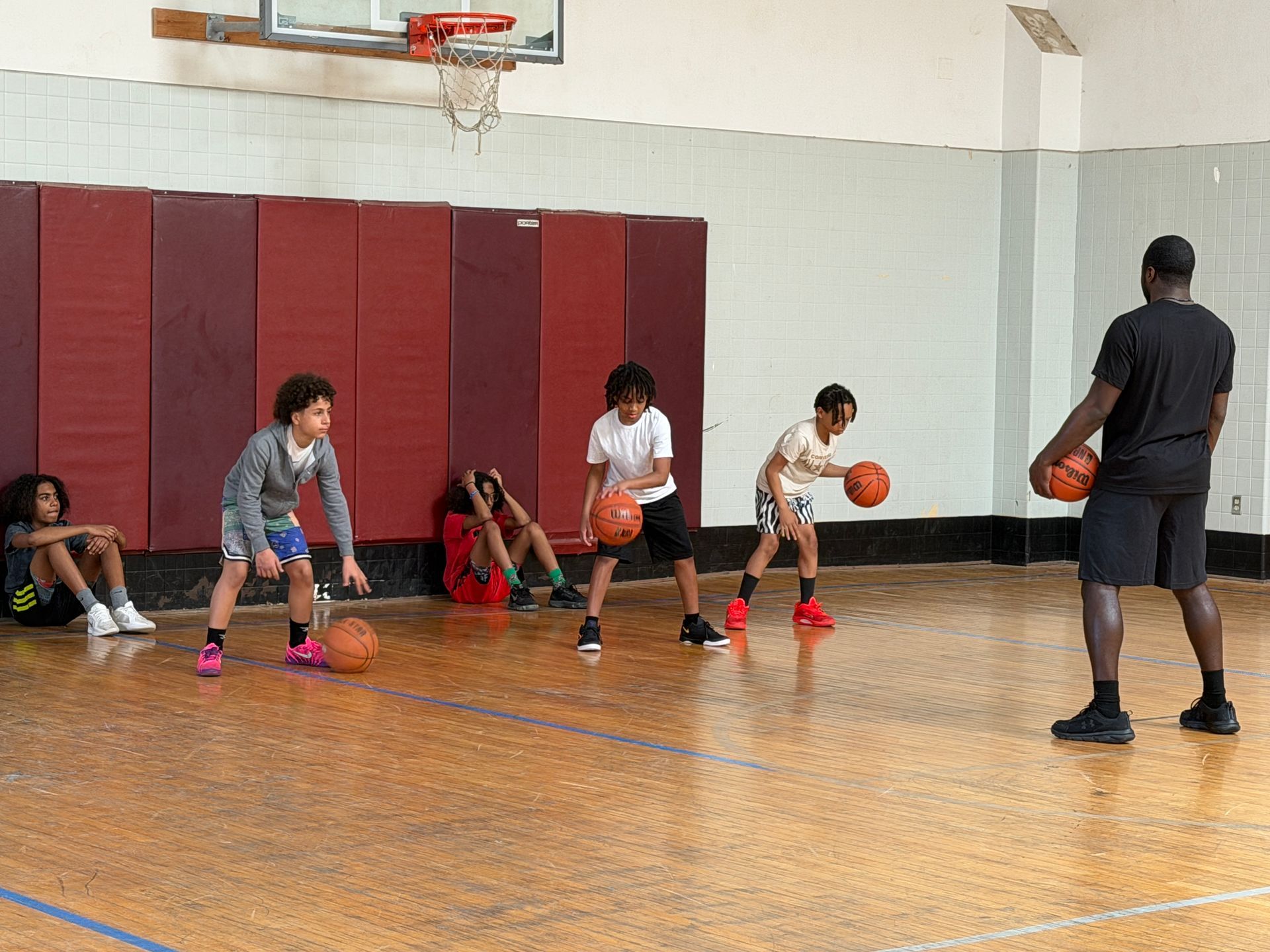 Kids dribbling basketballs in a gym during a practice drill with a coach watching.