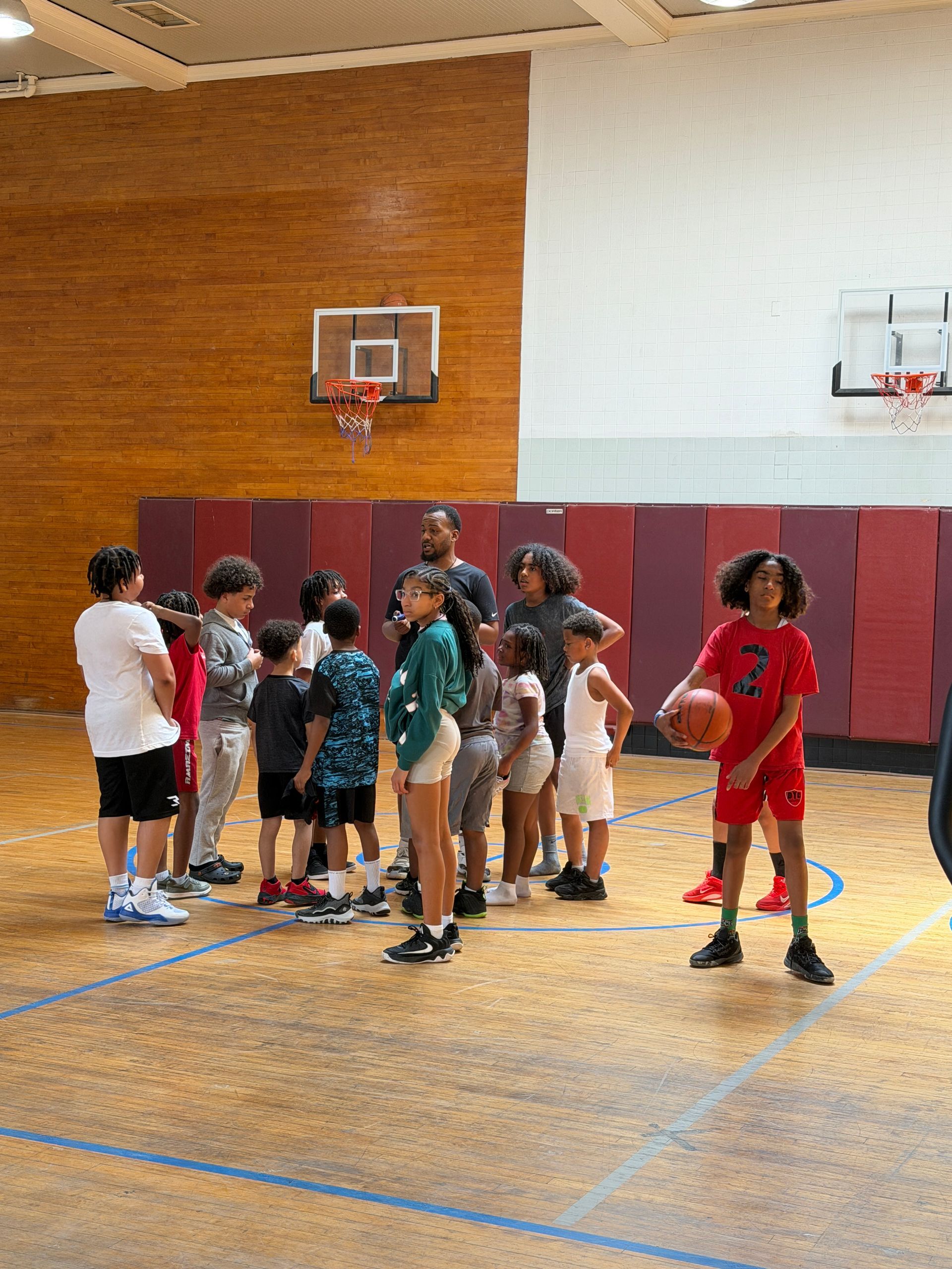 Children gathered on an indoor basketball court, with one in red holding a ball near the hoop.