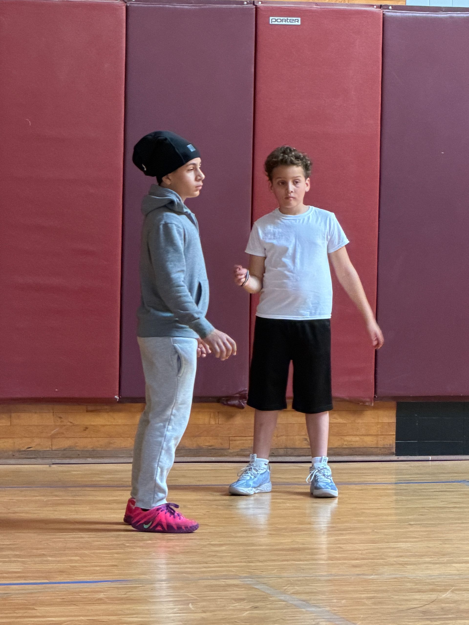 Two people standing on a gym floor in front of maroon wall padding, facing each other.