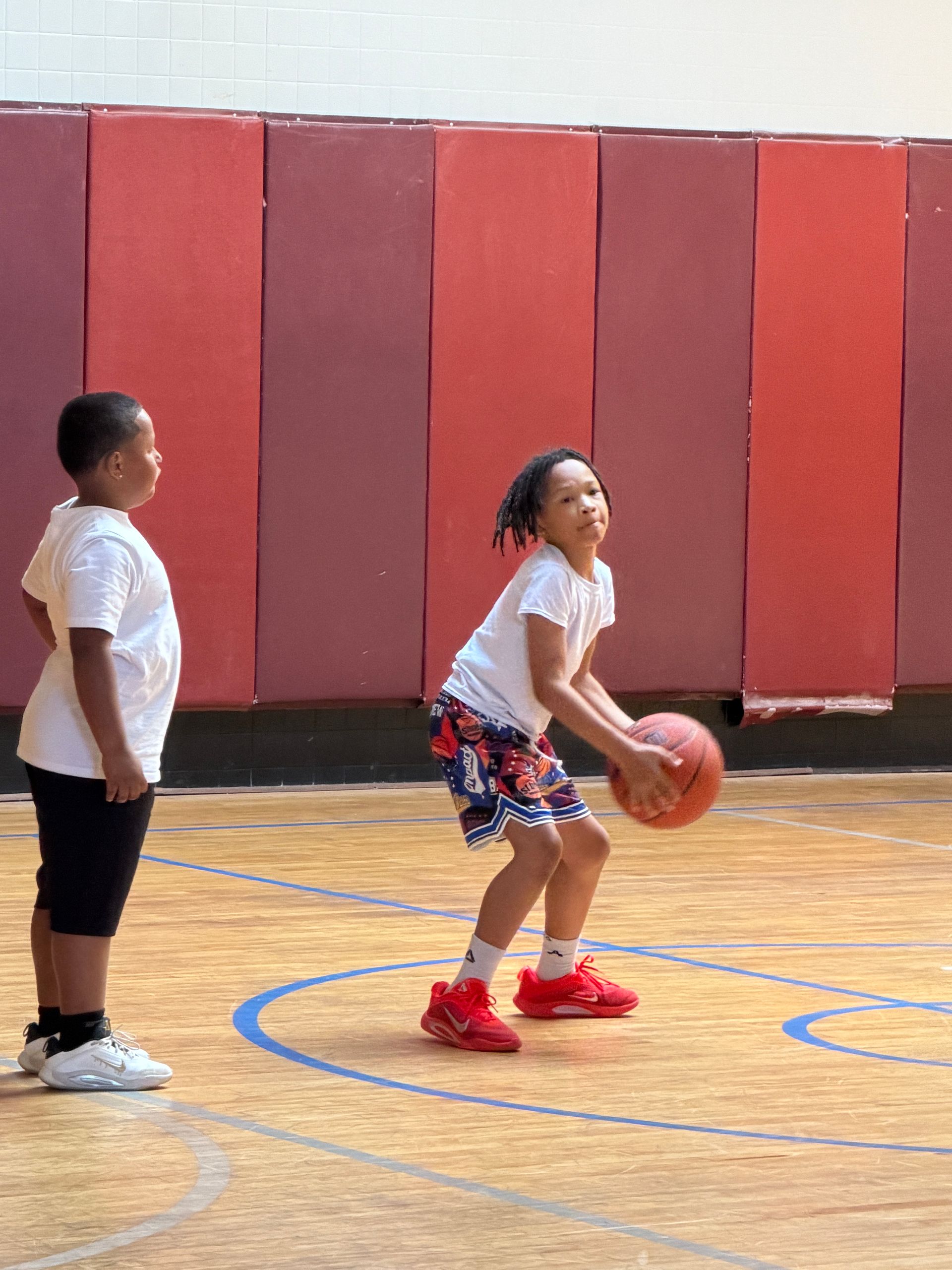 Two children on an indoor basketball court, one dribbling a ball while the other waits nearby