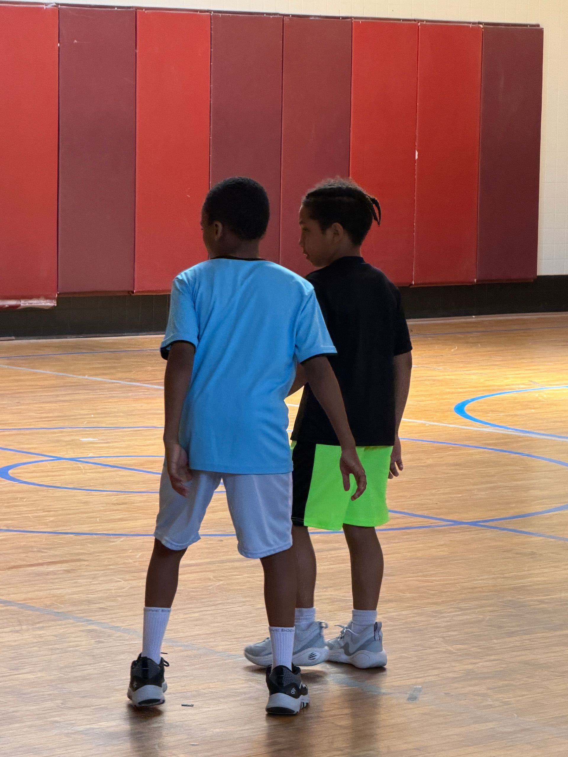 Two children standing on an indoor basketball court, facing each other near red wall padding.