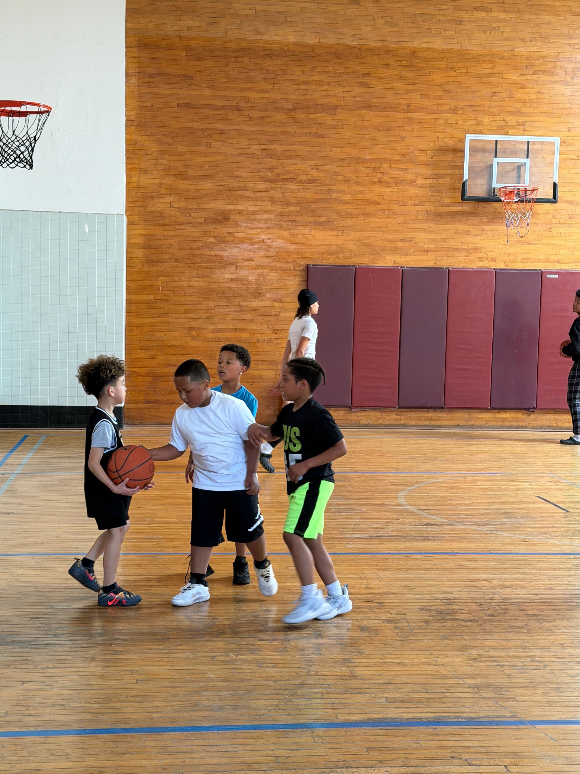 Children playing basketball in a gym, with one dribbling and others watching near the court.