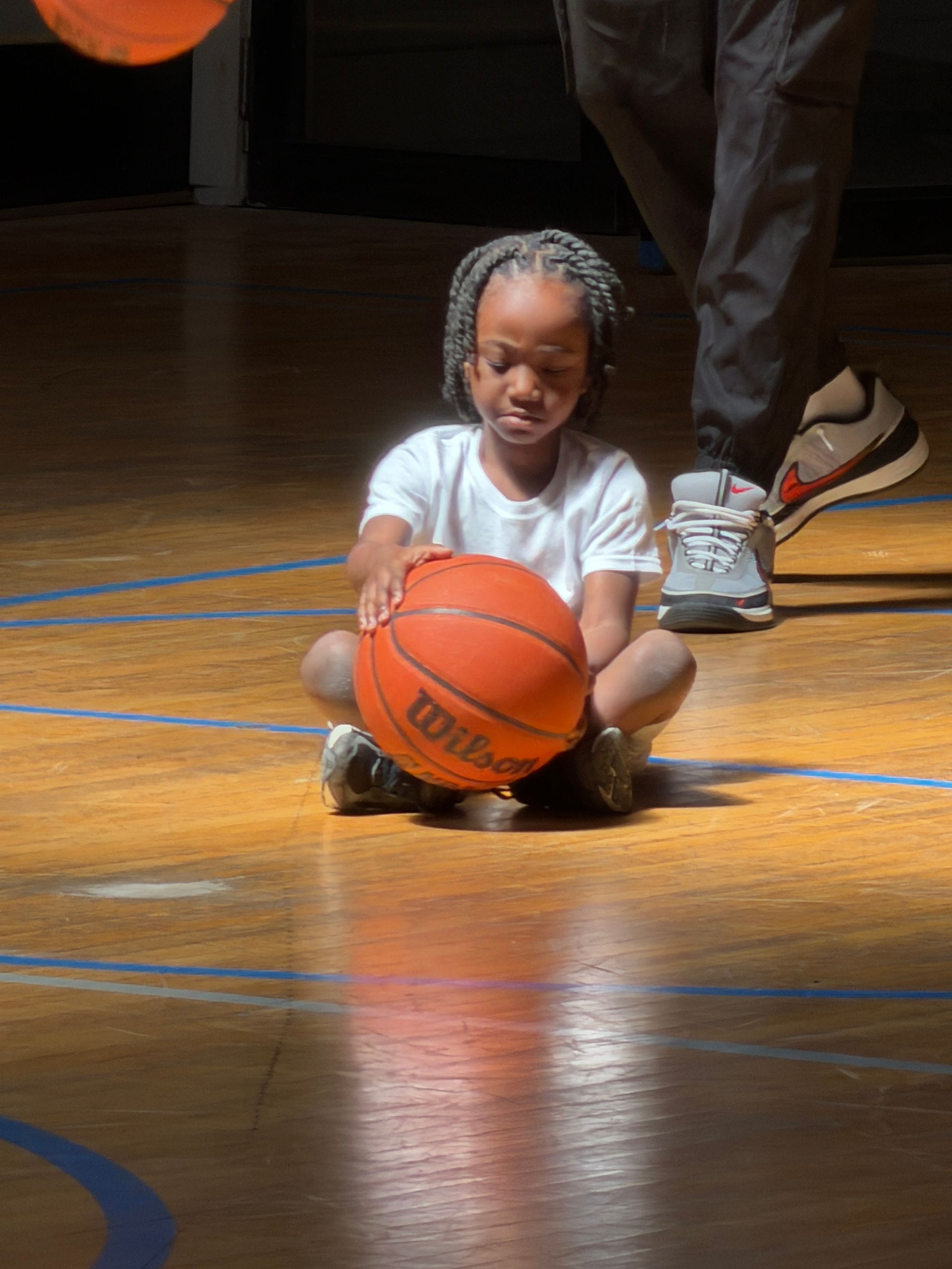 Child sitting on a gym floor holding an orange basketball, with an adult nearby