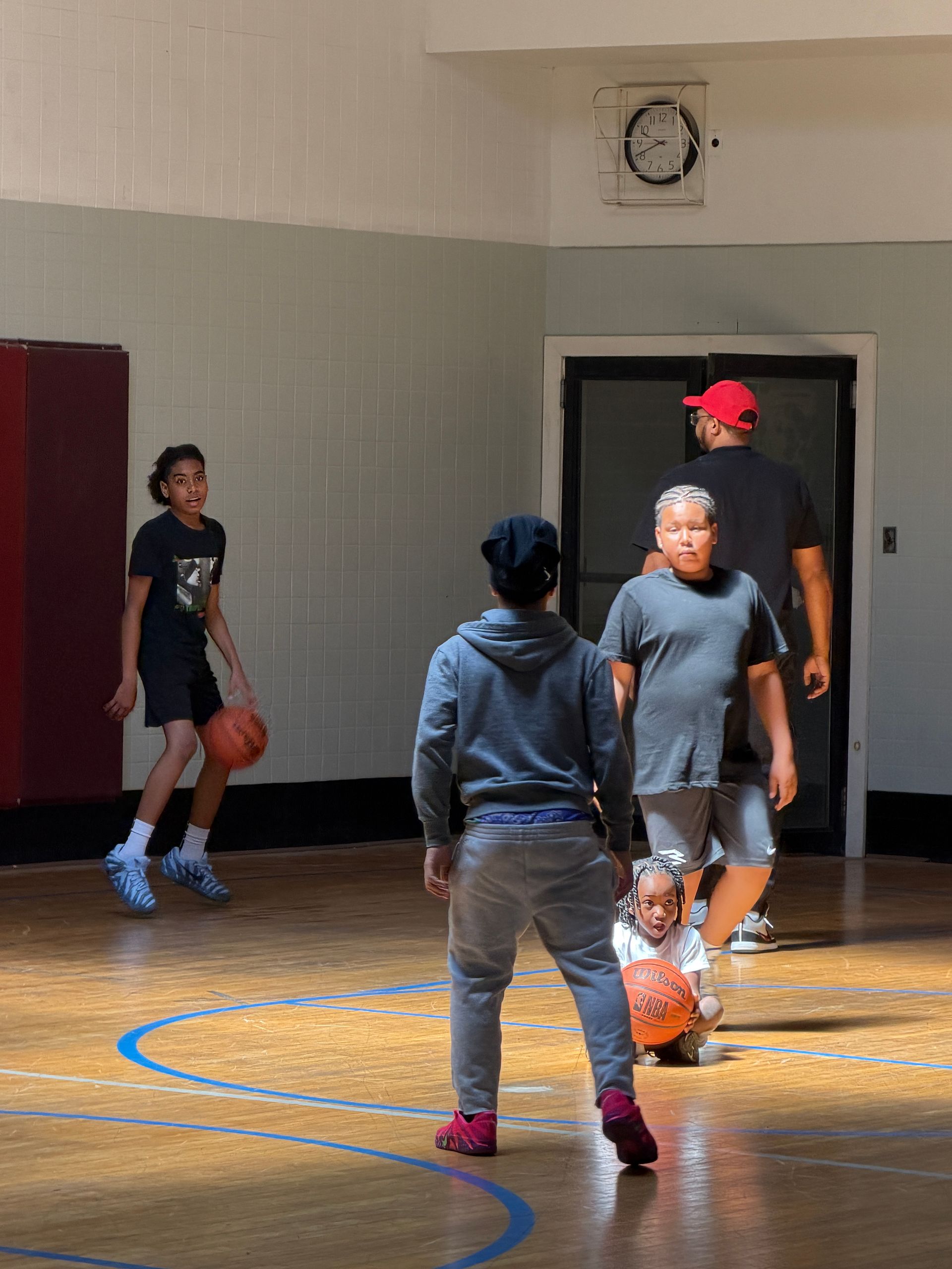 People playing basketball indoors, with one player dribbling near a hoop and others nearby.