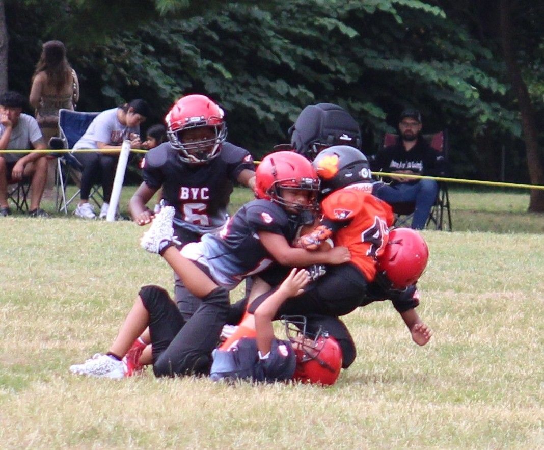 Youth tackle football players in red and black helmets pile up on a grassy field during a game.