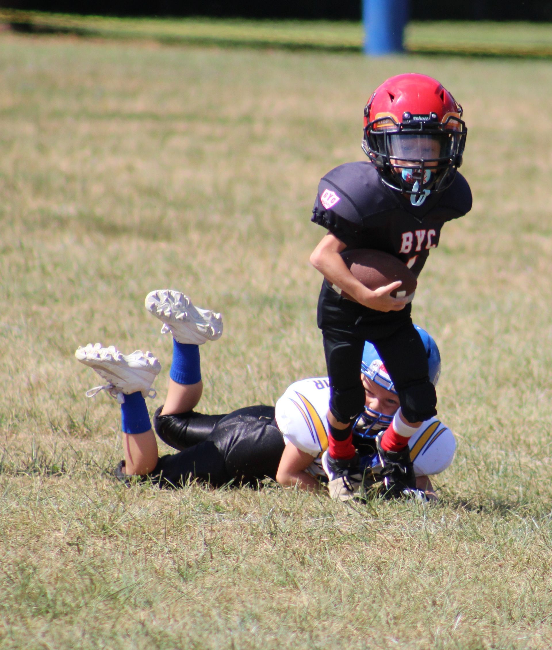Two children in football uniforms on a grassy field, one standing over another on the ground.