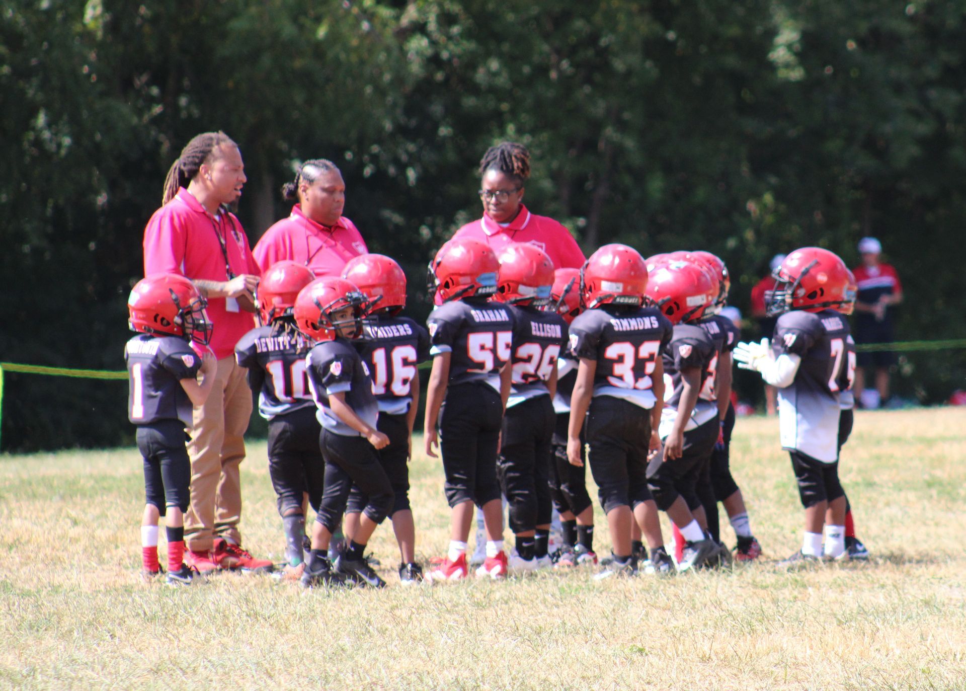 Youth football team huddled on a grassy field with coaches in red jerseys.