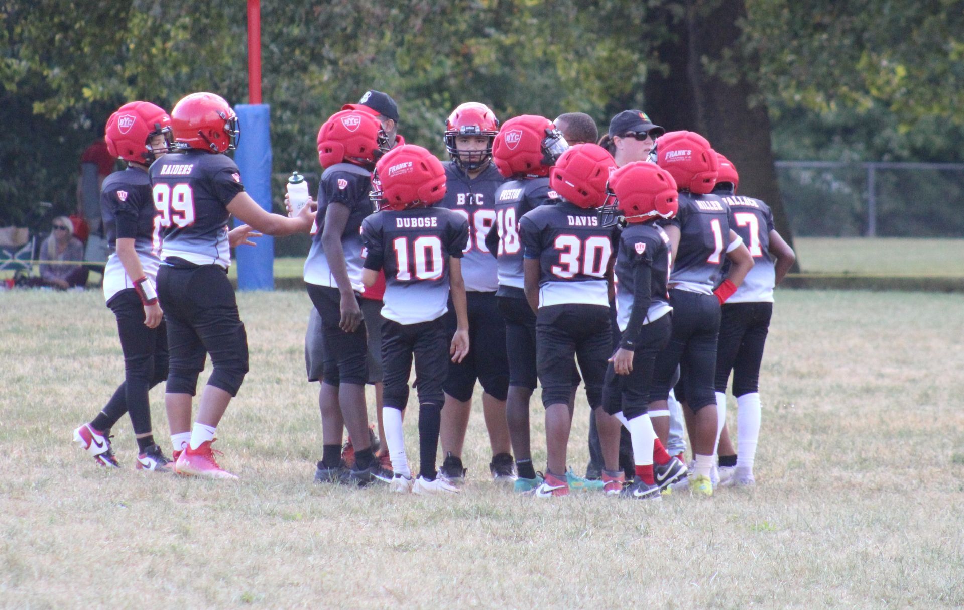 Youth football team in red helmets huddles on a grassy field near a goalpost.