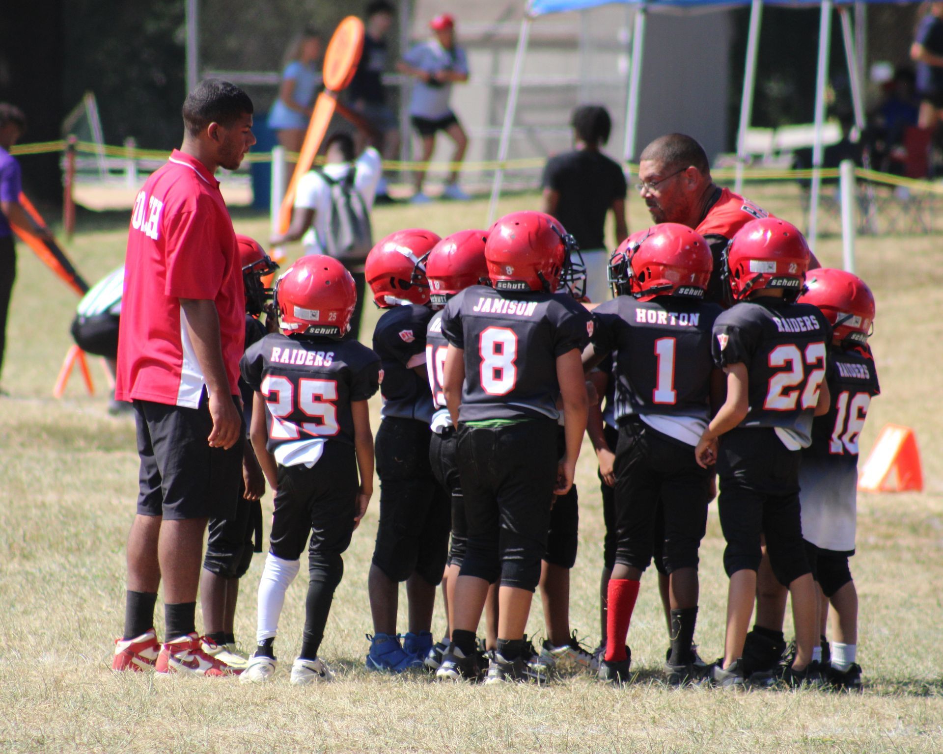 Youth football team in red helmets huddles with coaches on a grassy field during a game break