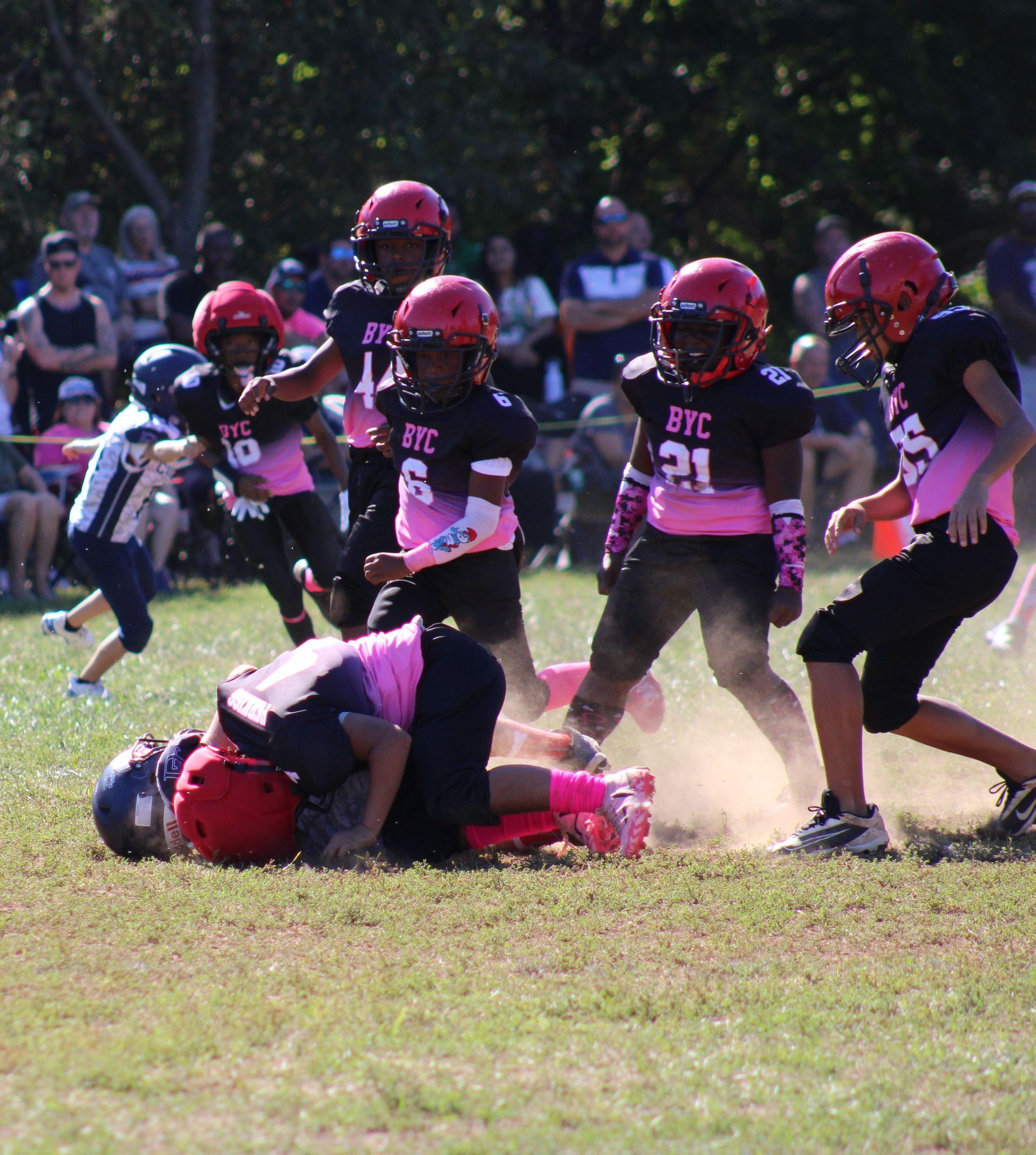 Youth football players in red and black tackle on a grassy field, with dust kicking up behind them.