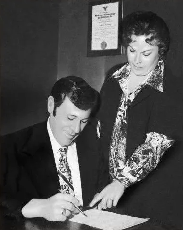 Man signing a document while a woman points to the page beside him in an office setting
