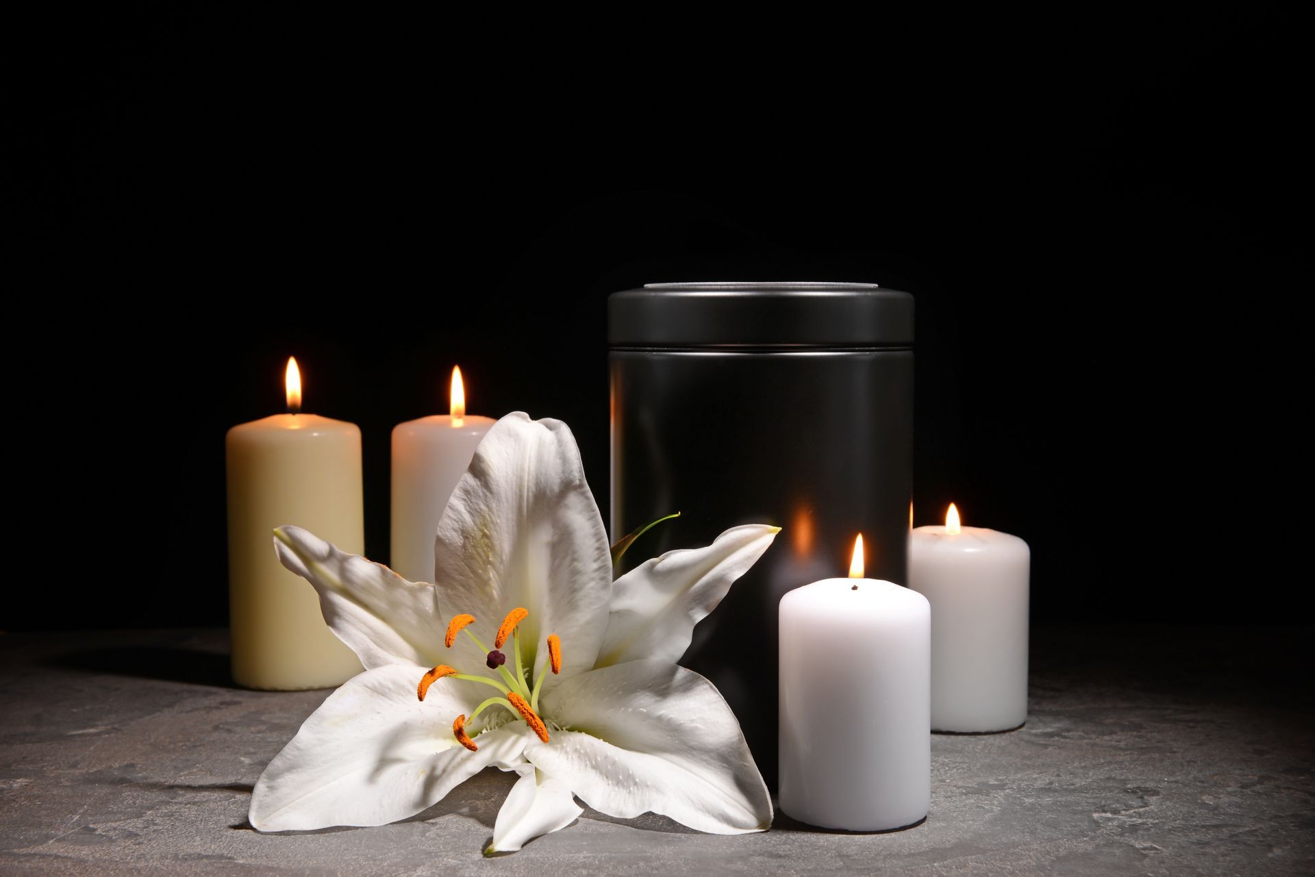 White lilies and lit candles beside a black urn on a dark table, creating a somber memorial scene