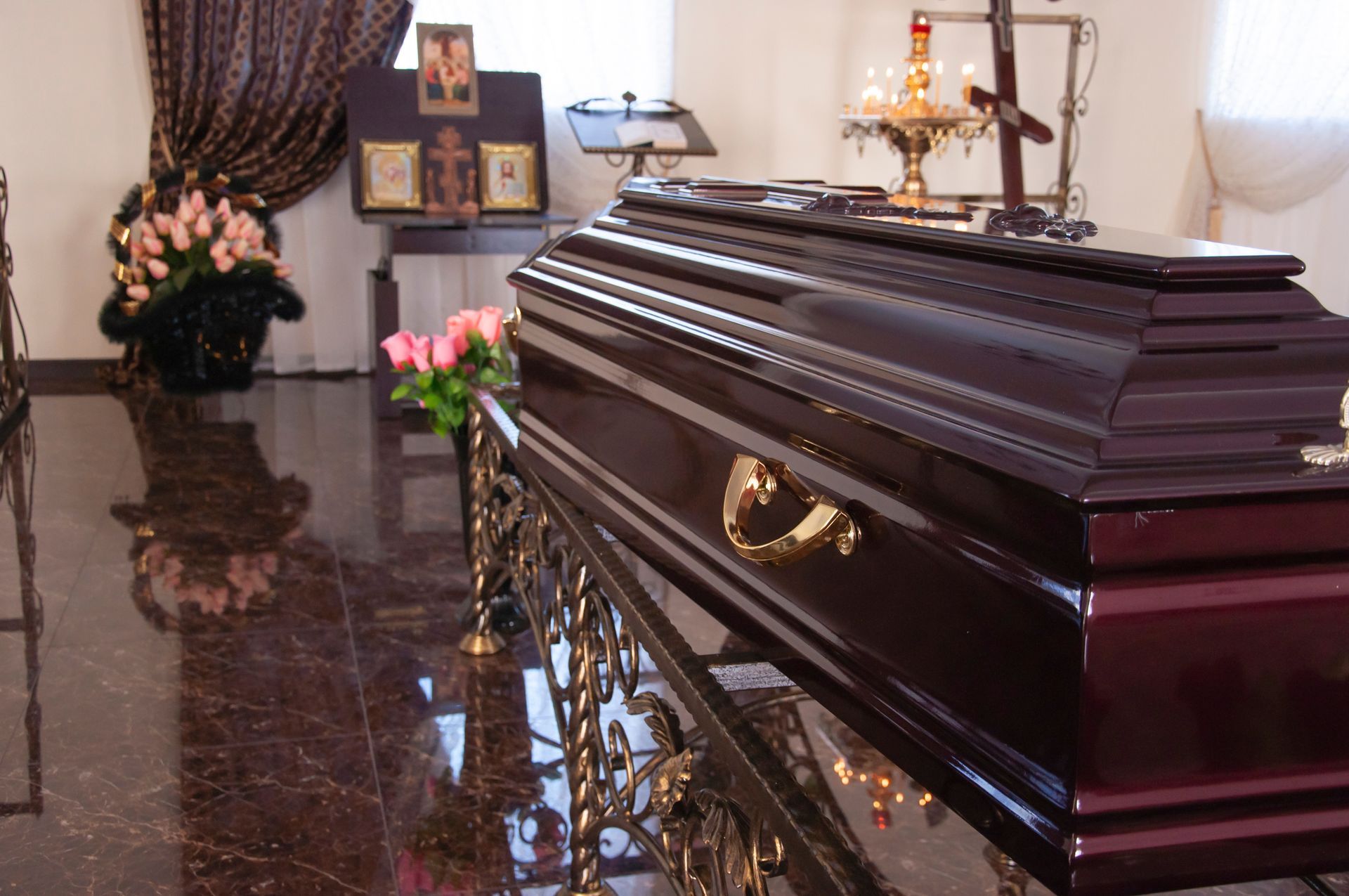 Brown coffin with gold handles in a funeral parlor, surrounded by flowers and a candlelit casket stand.