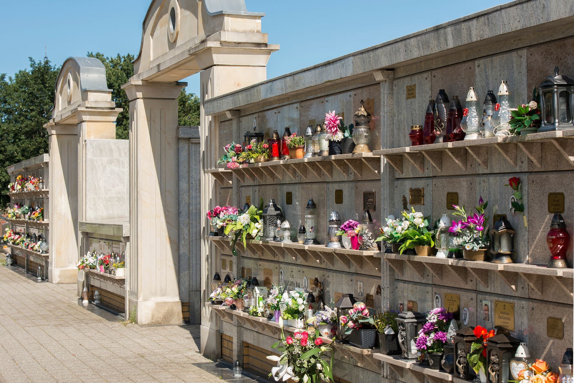 Outdoor cemetery niches lined with colorful flower arrangements along a stone wall