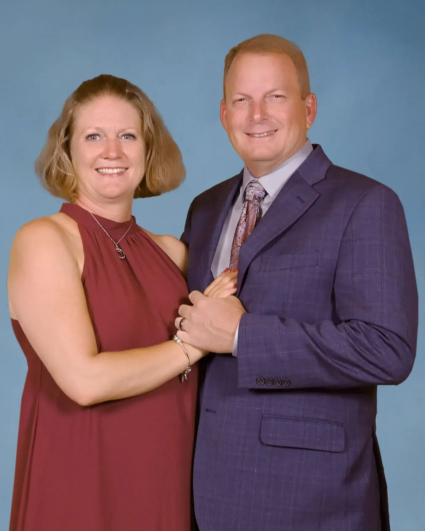 Two people posing together against a blue studio backdrop, smiling and holding hands.
