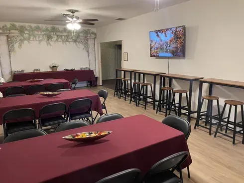 Empty banquet hall with burgundy tablecloths, black chairs, bar stools, and a wall-mounted TV.